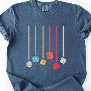 May include: A blue t-shirt featuring a graphic of colorful dice hanging from vertical lines. The dice are in various shapes and colors, including white, yellow, red, and blue. The shirt is made of a soft material and has a classic crew neck.