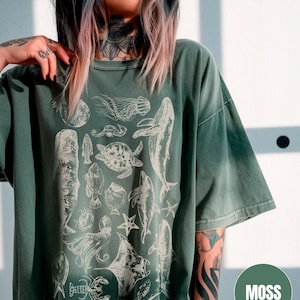 May include: A green oversized t-shirt with a white print of various sea creatures, including a whale, a turtle, a jellyfish, and a crab. The text "MOSS" is visible in the lower right corner of the image.