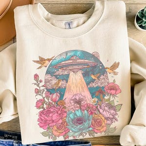 May include: Cream-colored sweatshirt featuring a colorful graphic of a UFO beaming light onto a floral arrangement with birds. The design includes pink, blue, and orange flowers. The sweatshirt is paired with blue denim shorts and a brown hat.