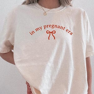 May include: Ivory-colored t-shirt with the phrase "in my pregnant era" in red script and a red bow graphic. The shirt is paired with red and white checkered shorts. The overall style is casual and comfortable.
