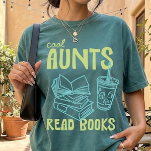 May include: A teal t-shirt with the text "cool AUNTS READ BOOKS" in a lime green font. The graphic includes a stack of books and a beverage cup. The shirt is paired with a white pleated skirt.