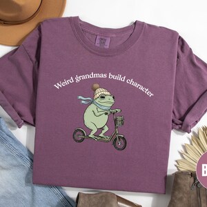 May include: Berry-colored t-shirt with rolled sleeves. The shirt features a whimsical illustration of a frog riding a scooter, wearing a hat and scarf. Text above the frog reads "Weird grandmas build character."