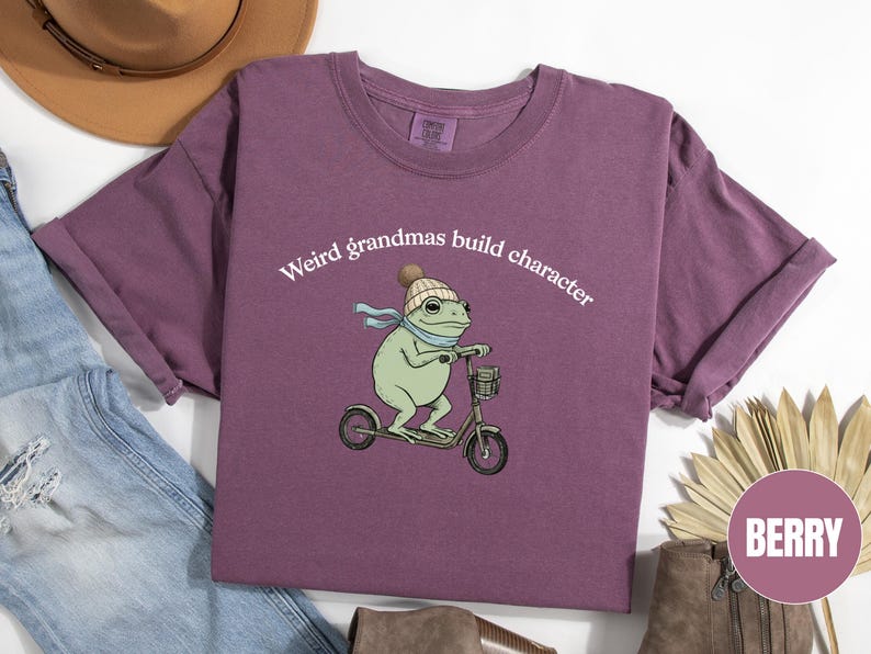May include: Berry-colored t-shirt with rolled sleeves. The shirt features a whimsical illustration of a frog riding a scooter, wearing a hat and scarf. Text above the frog reads "Weird grandmas build character."