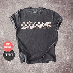 May include: Dark gray t-shirt featuring a playful design of cartoon ghosts on a checkered pattern. The ghosts are white with black accents, and the design includes autumn leaves. The shirt has rolled-up sleeves.