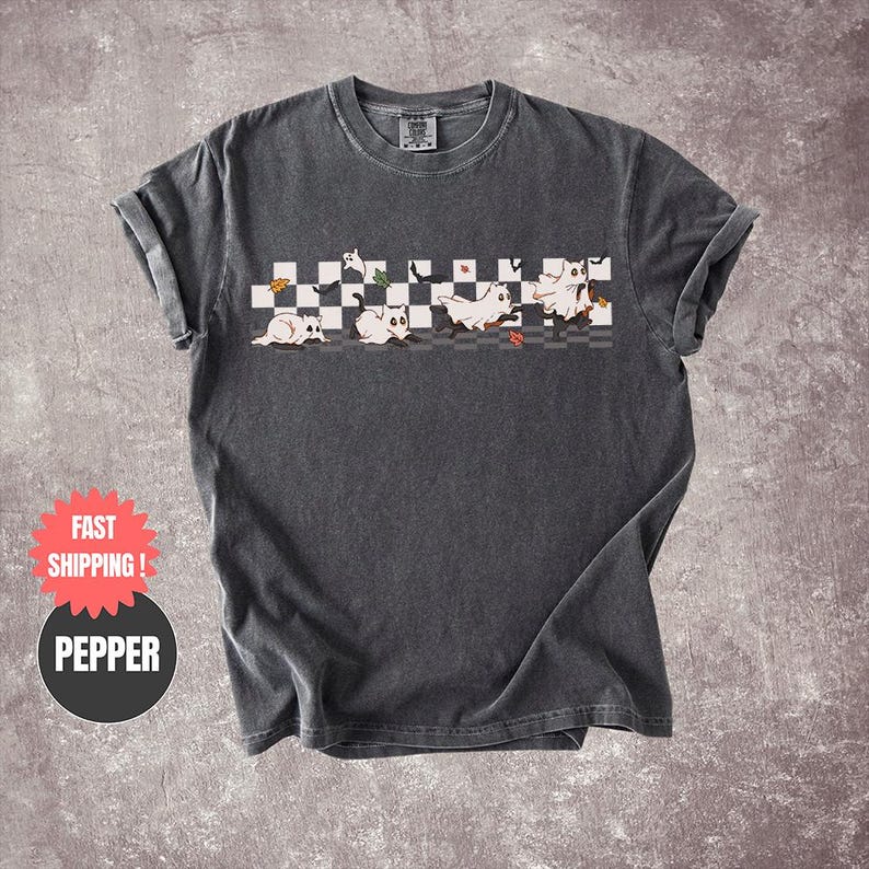 May include: Dark gray t-shirt featuring a playful design of cartoon ghosts on a checkered pattern. The ghosts are white with black accents, and the design includes autumn leaves. The shirt has rolled-up sleeves.