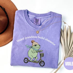 May include: A violet-colored t-shirt with the text "Weird grandmas build character." The shirt features a cartoon frog wearing a hat and scarf, riding a scooter. A gold necklace and a brown hat are also visible.