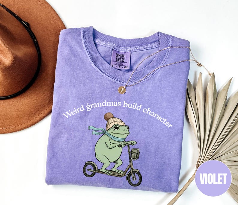 May include: A violet-colored t-shirt with the text "Weird grandmas build character." The shirt features a cartoon frog wearing a hat and scarf, riding a scooter. A gold necklace and a brown hat are also visible.