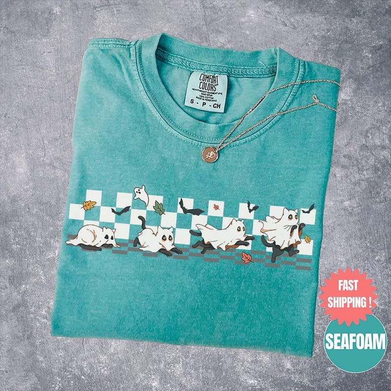 May include: Seafoam green t-shirt with a graphic of cats dressed as ghosts running across a checkered pattern. The shirt has a crew neck and a small tag at the neckline. A gold necklace with a pendant is draped across the shirt.