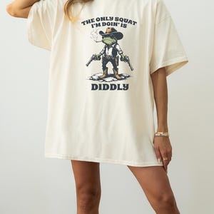 May include: Cream-colored t-shirt with the text "THE ONLY SQUAT I'M DOIN' IS DIDDLY". The graphic features a frog wearing a cowboy hat, smoking a cigarette, and holding two guns. The shirt has short sleeves and a relaxed fit.