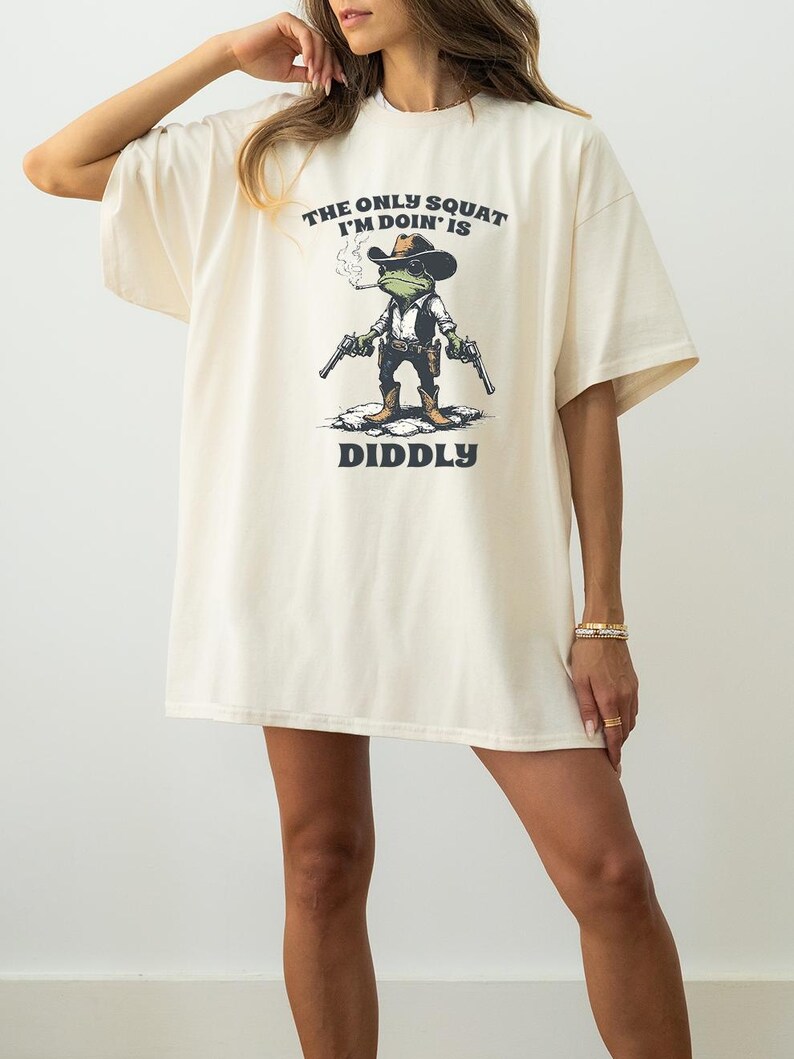May include: Cream-colored t-shirt with the text "THE ONLY SQUAT I'M DOIN' IS DIDDLY". The graphic features a frog wearing a cowboy hat, smoking a cigarette, and holding two guns. The shirt has short sleeves and a relaxed fit.