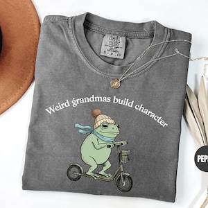 May include: A gray t-shirt with the text "Weird grandmas build character" above a cartoon frog riding a scooter. The frog is green with a blue scarf and a knit cap. The shirt has a crew neck.