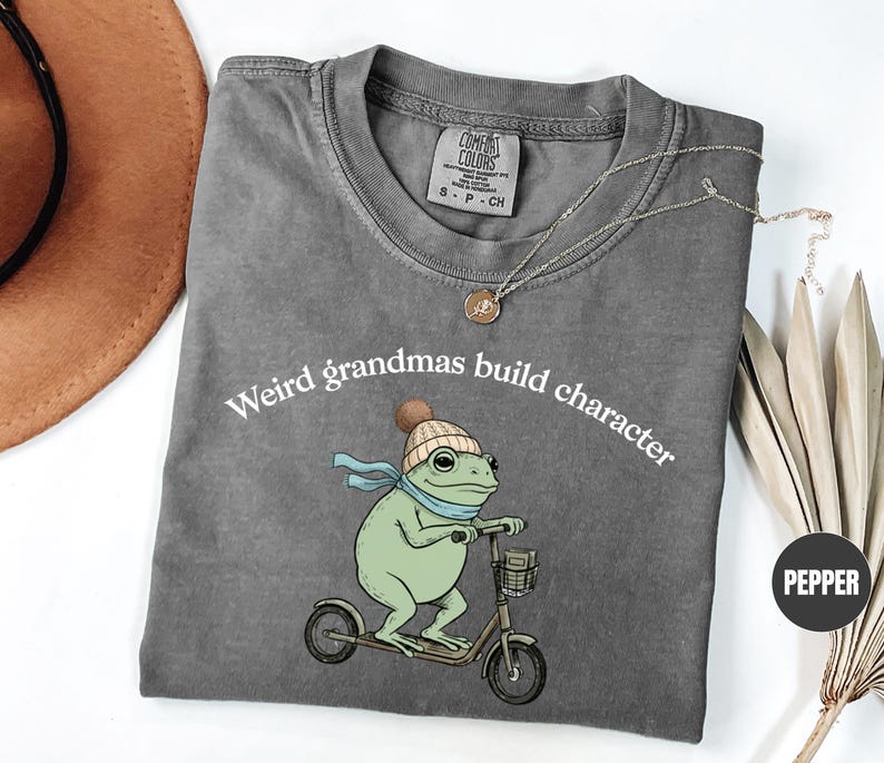 May include: A gray t-shirt with the text "Weird grandmas build character" above a cartoon frog riding a scooter. The frog is green with a blue scarf and a knit cap. The shirt has a crew neck.