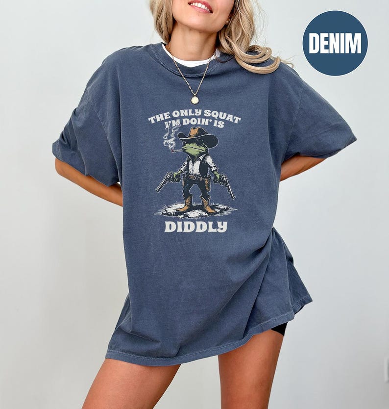 May include: Denim blue t-shirt featuring a cartoon frog dressed as a cowboy, holding two guns and smoking a cigarette. The text on the shirt reads "THE ONLY SQUAT I'M DOIN' IS DIDDLY".