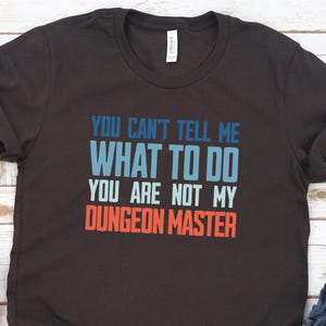 Puede incluir: Una camiseta gris oscuro con el texto "You can't tell me what to do. You are not my Dungeon Master." en azul, blanco y naranja.