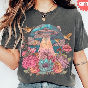 May include: Dark grey t-shirt with a vibrant graphic of a UFO shining a beam of light onto flowers. The design includes birds, clouds, and a blue and pink sky. The shirt has a vintage, faded appearance.