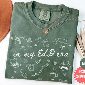 May include: A green Comfort Colors t-shirt with the phrase "in my EdD era" and various white graphic icons. The shirt has a crew neck and short sleeves. A gold necklace with a pendant is draped on the shirt.
