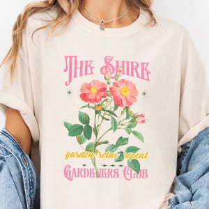 May include: A beige t-shirt with a pink graphic design that says "The Shire Garden Relax Repeat Gardeners Club". The graphic features pink roses and green leaves.