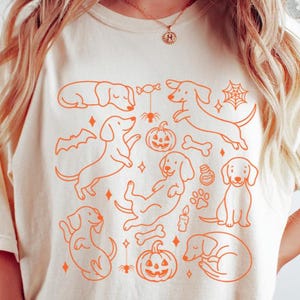 May include: Cream-colored t-shirt featuring an orange outline design of dachshund dogs, pumpkins, bats, spiderwebs, and bones. The design has a Halloween theme.