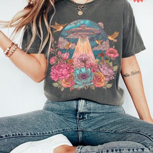 May include: Dark grey t-shirt with a colourful graphic of a UFO beaming light onto flowers and birds. The shirt is worn with blue jeans and white trainers. The image has a fast shipping tag.