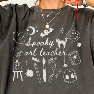 May include: Dark gray sweatshirt with the text "Spooky art teacher" and white illustrations of a ghost, bat, cat, paint palette, and skull. The shirt has a crew neck and long sleeves.