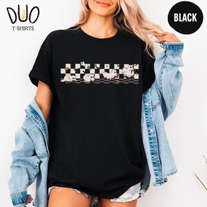 May include: Black t-shirt with a graphic of cartoon cats on a checkered pattern. The shirt has short sleeves and a crew neck. The brand logo "DUO T-SHIRTS" is visible in the upper left corner. A black circle with the word "BLACK" is in the upper right corner.