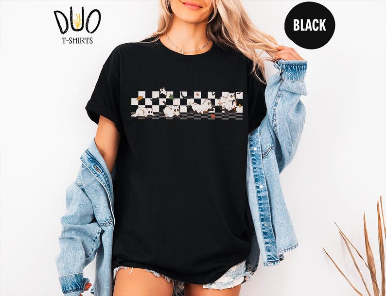 May include: Black t-shirt with a graphic of cartoon cats on a checkered pattern. The shirt has short sleeves and a crew neck. The brand logo "DUO T-SHIRTS" is visible in the upper left corner. A black circle with the word "BLACK" is in the upper right corner.