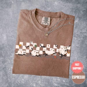 May include: Espresso-colored t-shirt with a playful Halloween design. The graphic features cartoon cats dressed as ghosts running across a checkered pattern, with falling leaves and bats. A silver necklace with a pendant rests on the shirt.