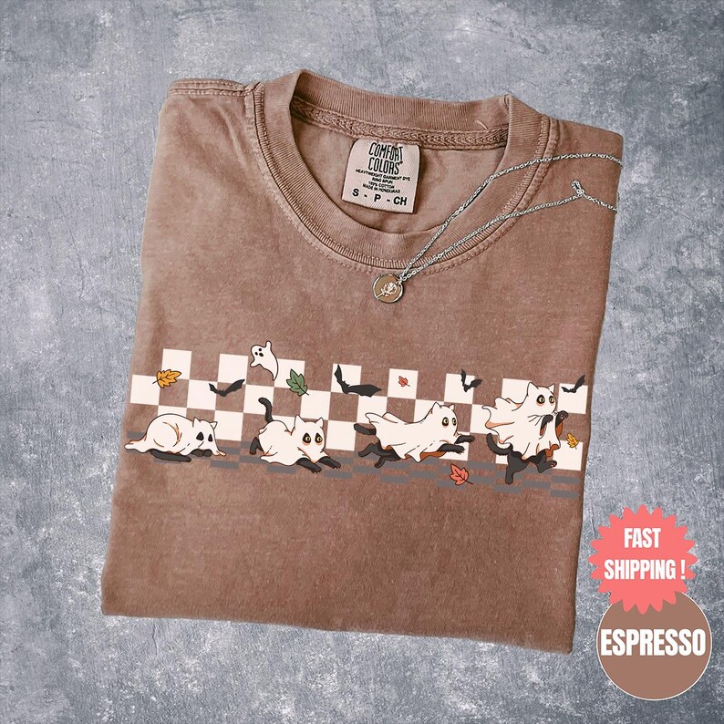 May include: Espresso-colored t-shirt with a playful Halloween design. The graphic features cartoon cats dressed as ghosts running across a checkered pattern, with falling leaves and bats. A silver necklace with a pendant rests on the shirt.