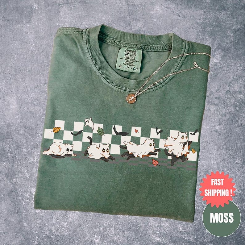 May include: A moss green t-shirt with a graphic of cats dressed as ghosts running across a checkered pattern. The shirt has a crew neck and a silver necklace with a pendant. The shirt is folded and has a tag that says "Comfort Colors".