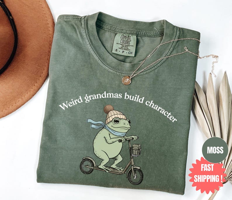 May include: A moss green t-shirt with the text "Weird grandmas build character" and a cartoon frog riding a scooter. The frog wears a knit cap and scarf. A gold necklace and a brown hat are also visible.