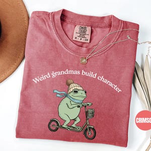 May include: Crimson-colored t-shirt with the text "Weird grandmas build character." The shirt features a cartoon frog wearing a hat and scarf, riding a scooter. A gold necklace is draped on the shirt.