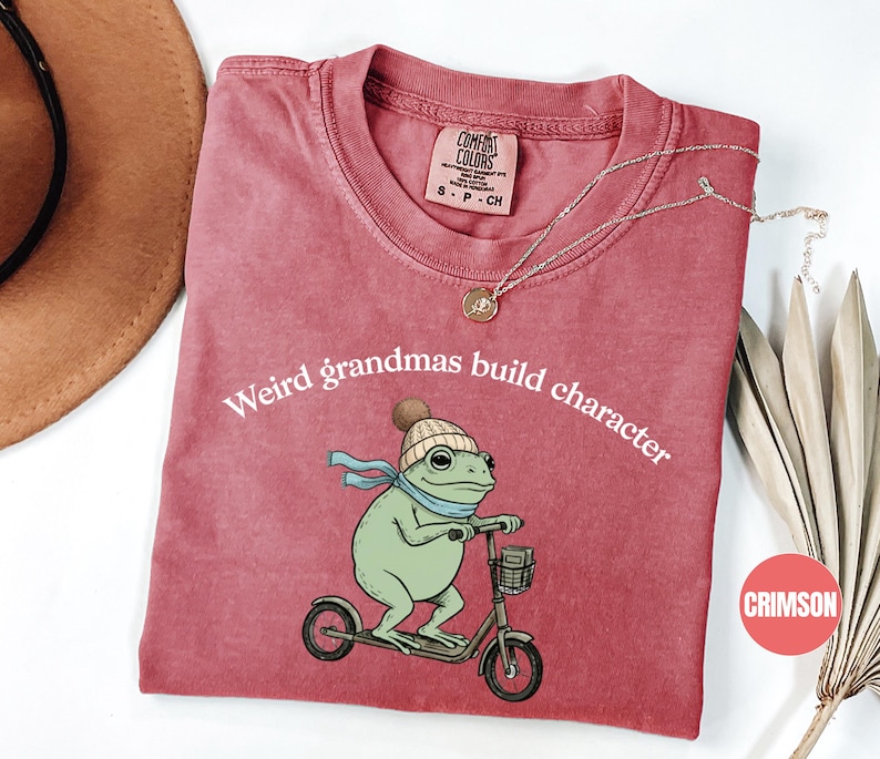 May include: Crimson-colored t-shirt with the text "Weird grandmas build character." The shirt features a cartoon frog wearing a hat and scarf, riding a scooter. A gold necklace is draped on the shirt.