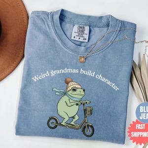 May include: A light blue t-shirt with the text "Weird grandmas build character" and a cartoon frog riding a scooter. The frog wears a hat and scarf. The shirt has a Comfort Colors tag.
