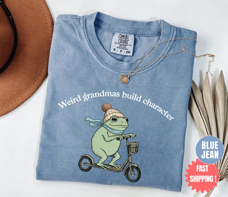 May include: A light blue t-shirt with the text "Weird grandmas build character" and a cartoon frog riding a scooter. The frog wears a hat and scarf. The shirt has a Comfort Colors tag.