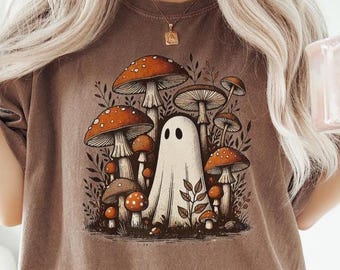 May include: Brown t-shirt featuring a whimsical illustration of a white ghost surrounded by orange and brown mushrooms. The design includes detailed botanical elements, creating a nature-inspired theme.