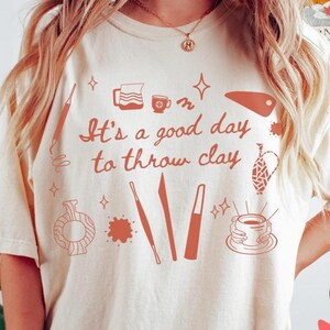 May include: Cream-colored t-shirt with a terracotta-colored design. The design includes pottery tools, a teapot, a cup, and the phrase "It's a good day to throw clay." The shirt has a crew neck and short sleeves.