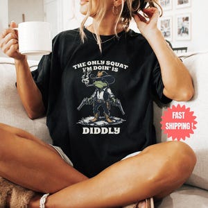 May include: Black t-shirt with a graphic of a frog cowboy holding two guns, a cigarette, and wearing a hat. The text reads "THE ONLY SQUAT I'M DOIN' IS DIDDLY". The shirt is worn by a person sitting on a couch.