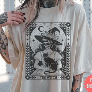 May include: Cream-colored t-shirt featuring a black and white illustration of a witch with a skull face, wearing a hat and holding a black cat. The text "THE WITCH" is printed at the bottom of the design, framed by decorative elements.