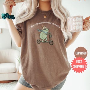 May include: A brown t-shirt with the text "Weird grandmas build character" above a cartoon frog riding a scooter. The frog wears a hat and scarf. A coffee mug is held in the hand.