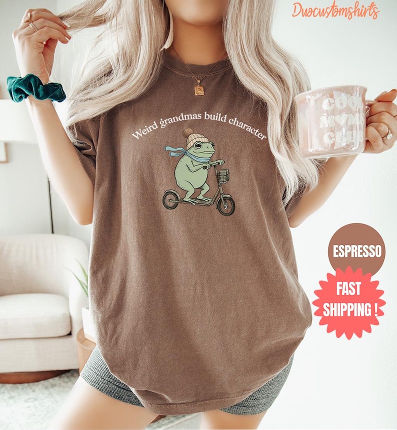 May include: A brown t-shirt with the text "Weird grandmas build character" above a cartoon frog riding a scooter. The frog wears a hat and scarf. A coffee mug is held in the hand.