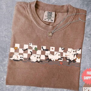 May include: Brown Comfort Colors t-shirt with a Halloween-themed graphic. The design features cartoon cats dressed as ghosts running across a checkered pattern. A silver necklace with a pendant rests on the shirt.