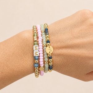 May include: A stack of beaded bracelets in various colors, including gold, pink, blue, and green. One bracelet features the letters "MM" on white square beads. Another has a gold charm with a bird design. The bracelets are worn on a wrist.