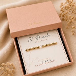 May include: A gold beaded bracelet with a single pearl bead, presented in a pink box. The box is open, revealing the bracelet on a card with the text "31 Beads" and "THE ESTHER". The card also includes the text "31 WOMEN. 1 STORY. EVERY SEASON."
