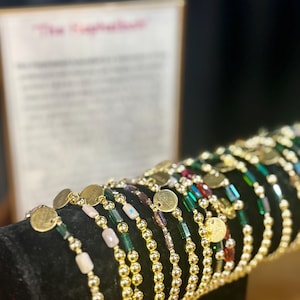 May include: A selection of beaded bracelets with gold-coloured details and small, round charms. The bracelets feature various bead colours, including green, pink, and clear, and are displayed on a black velvet stand.