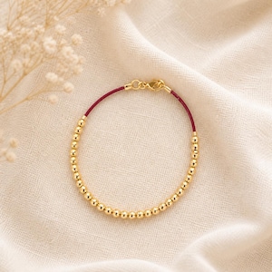 May include: Gold beaded bracelet with a burgundy cord and gold clasp. The bracelet is displayed on a cream-colored fabric, with dried flowers in the background. The bracelet is approximately 7 inches in length.