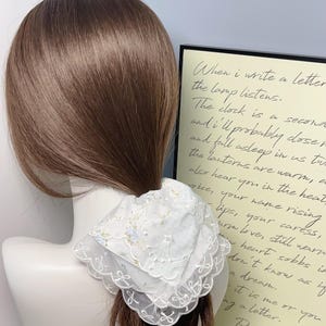 Puede incluir: Un scrunchie para el pelo de encaje blanco con un patrón floral. El scrunchie está atado alrededor de una coleta. El fondo es una pared con una cita sobre escribir cartas.