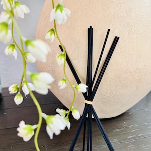 May include: A set of black diffuser sticks in a vase with white flowers.