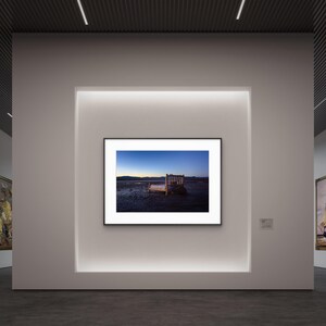 Op de afbeelding: Een ingelijste foto van een houten bank op een strand bij zonsondergang. De lucht gaat over van blauw naar oranje, met bergen in de verte. De foto wordt tentoongesteld in een galerie met witte muren en inbouwverlichting.