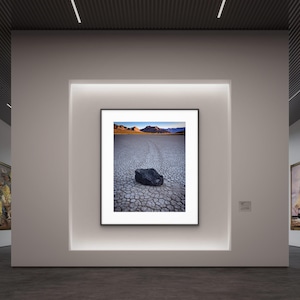 May include: A framed landscape photograph featuring a cracked earth surface with a dark rock in the foreground. The background shows a distant mountain range under a blue sky. The photograph is displayed in a gallery setting.