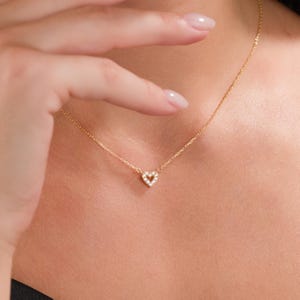 May include: A delicate gold necklace featuring a small heart pendant. The heart is encrusted with small, sparkling stones. The necklace is worn around the neck, with the chain visible. The overall design is simple and elegant.